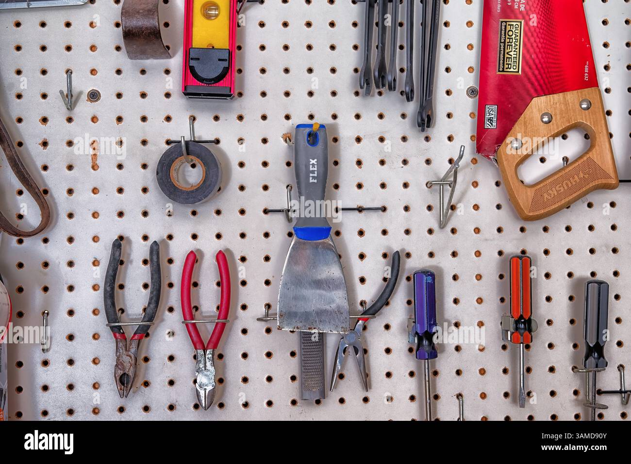 Eine Vielzahl von Handwerkzeugen wie Zangen, Schraubendreher und eine Säge, die auf einer weißen Wand in einer Werkstatt oder Garage angeordnet sind. Stockfoto