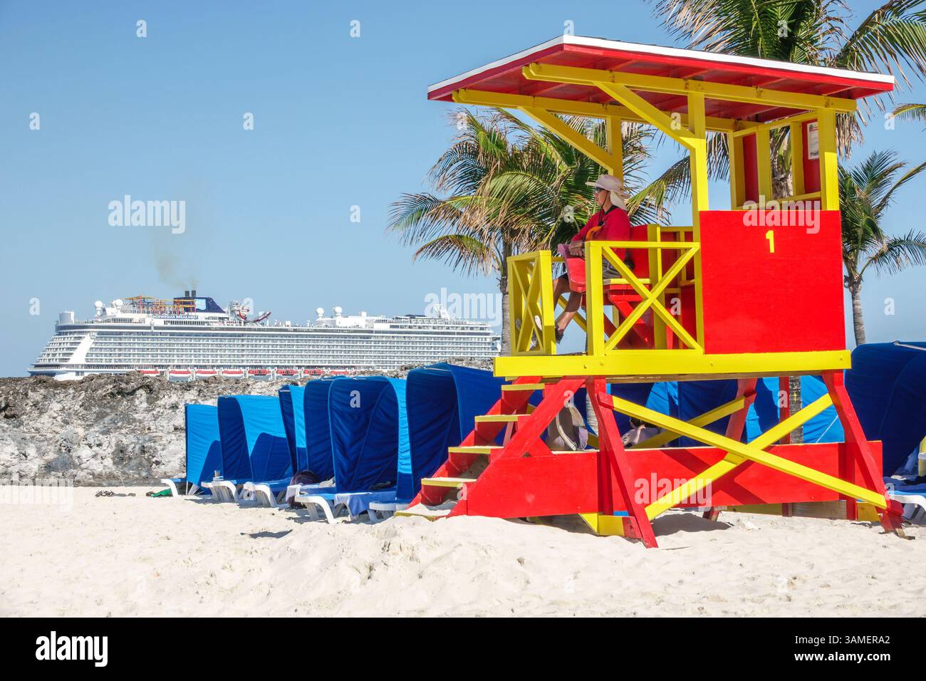Toller Stirrup Cay, die Bahamas, roter gelber Rettungsschwimmerturm, Kreuzfahrtschiff im Hintergrund, Strandhütten, blaue Liegestühle, weißer Sand, Palmen, tropischer isl Stockfoto