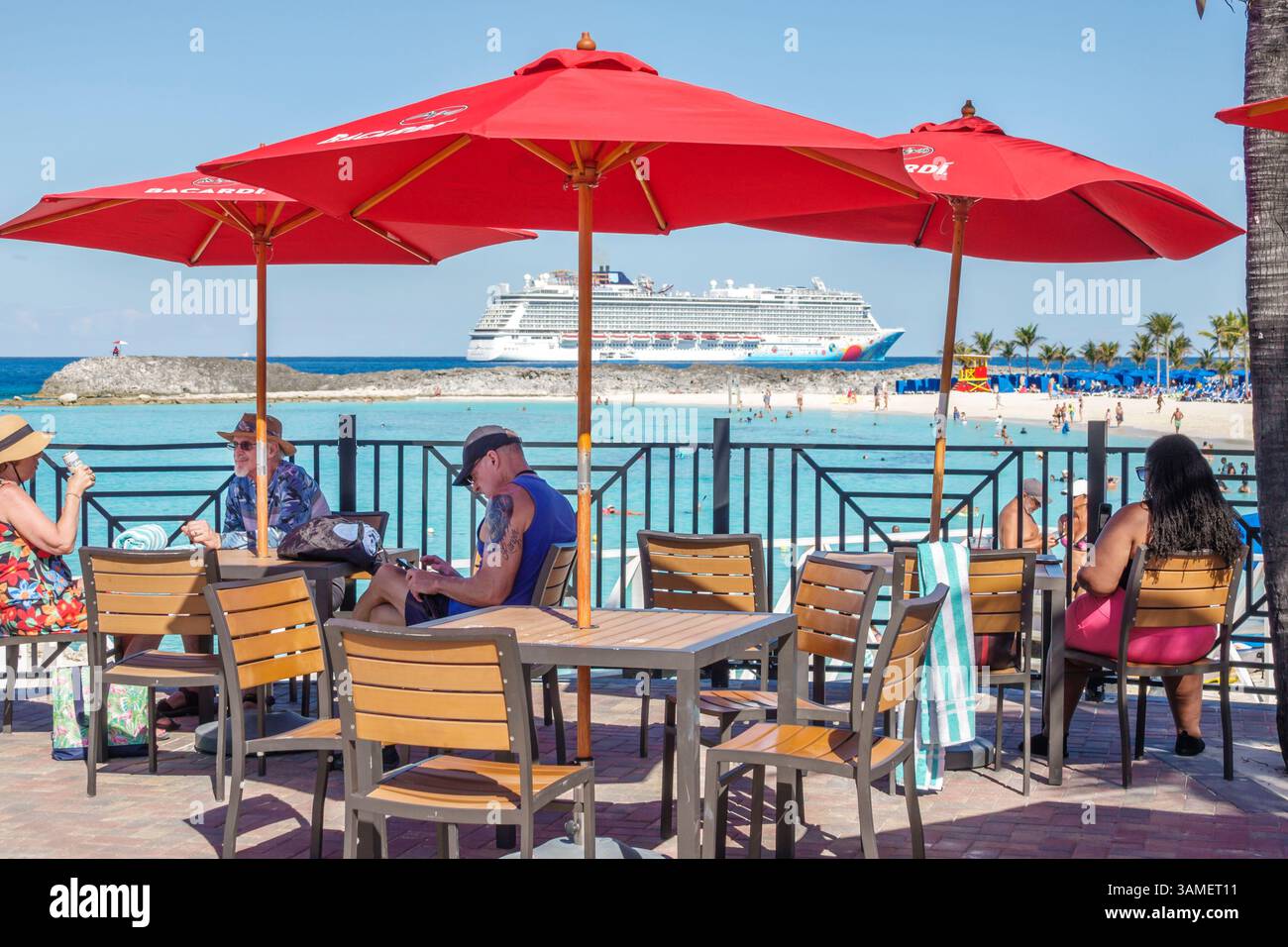 Großartiges Stirrup Cay, die Bahamas, am Strand mit roten Bacardi-Regenschirmen, Kreuzfahrtschiff vor der Küste vor Anker, Erwachsene sitzen entspannendes Resort-Café, Blick auf die Küste Stockfoto