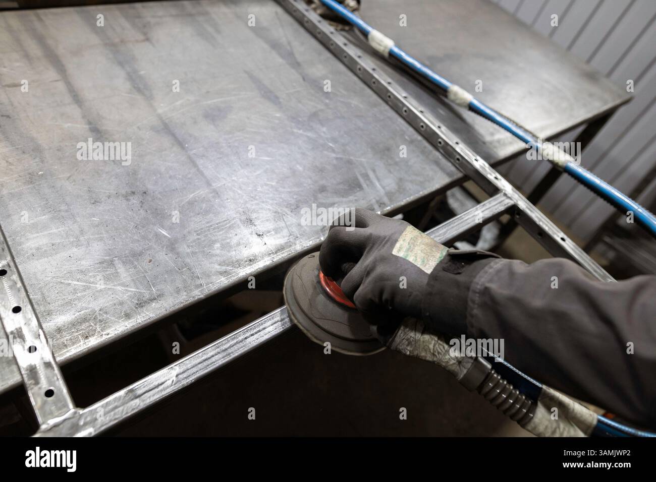 Die Handschuhe eines Arbeitnehmers verwenden einen Schleifmaschinen, um die Fugen eines Metallrahmens auf einer Werkbank in einer Werkstatt zu glätten. Stockfoto