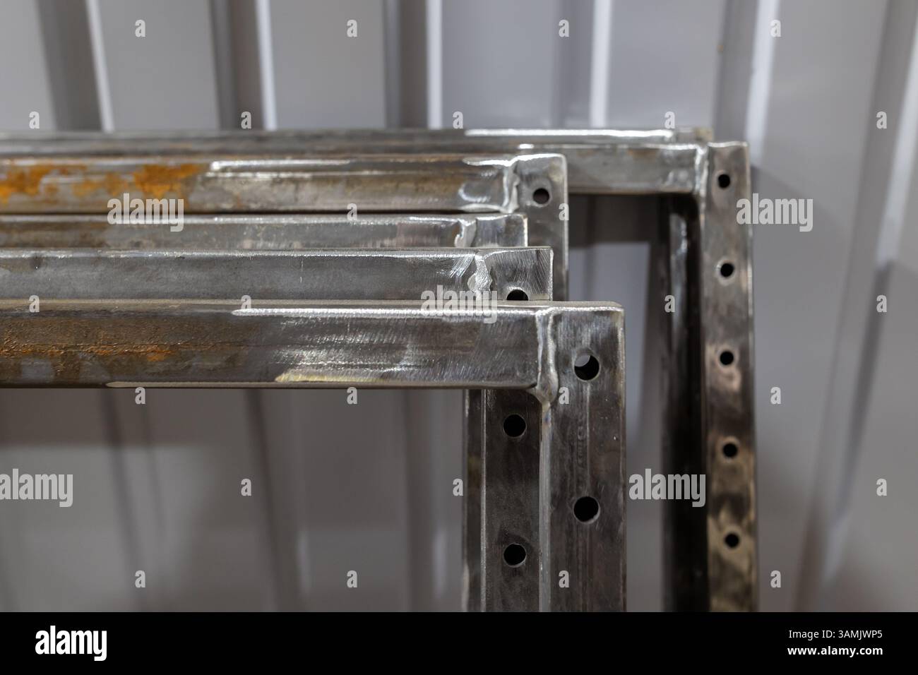 Mehrere geschweißte Metallrahmen mit gebohrten Löchern werden in einer Werkstattumgebung gegen eine gewellte Metallwand gestapelt. Stockfoto
