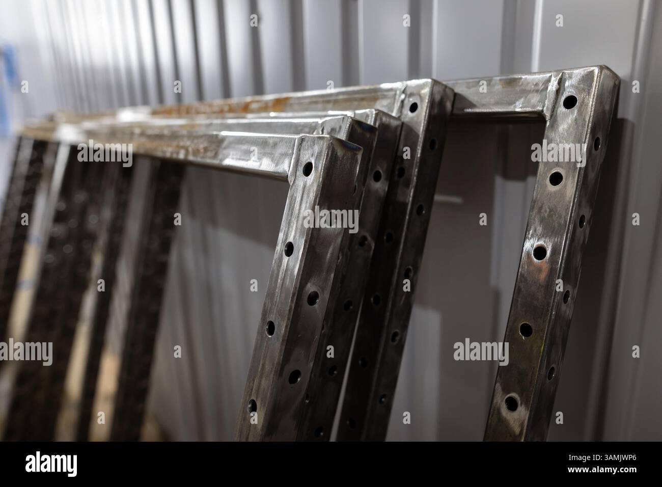 Mehrere geschweißte Metallrahmen mit gebohrten Löchern werden in einer Werkstattumgebung gegen eine gewellte Metallwand gestapelt. Stockfoto