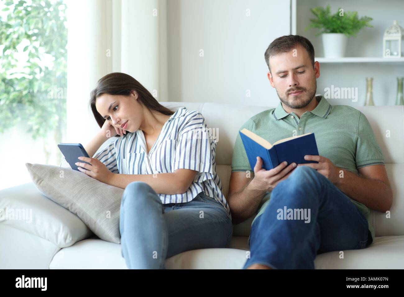 Ein Paar, das Papier liest und digitale Bücher liest, sitzt zu Hause auf einer Couch Stockfoto