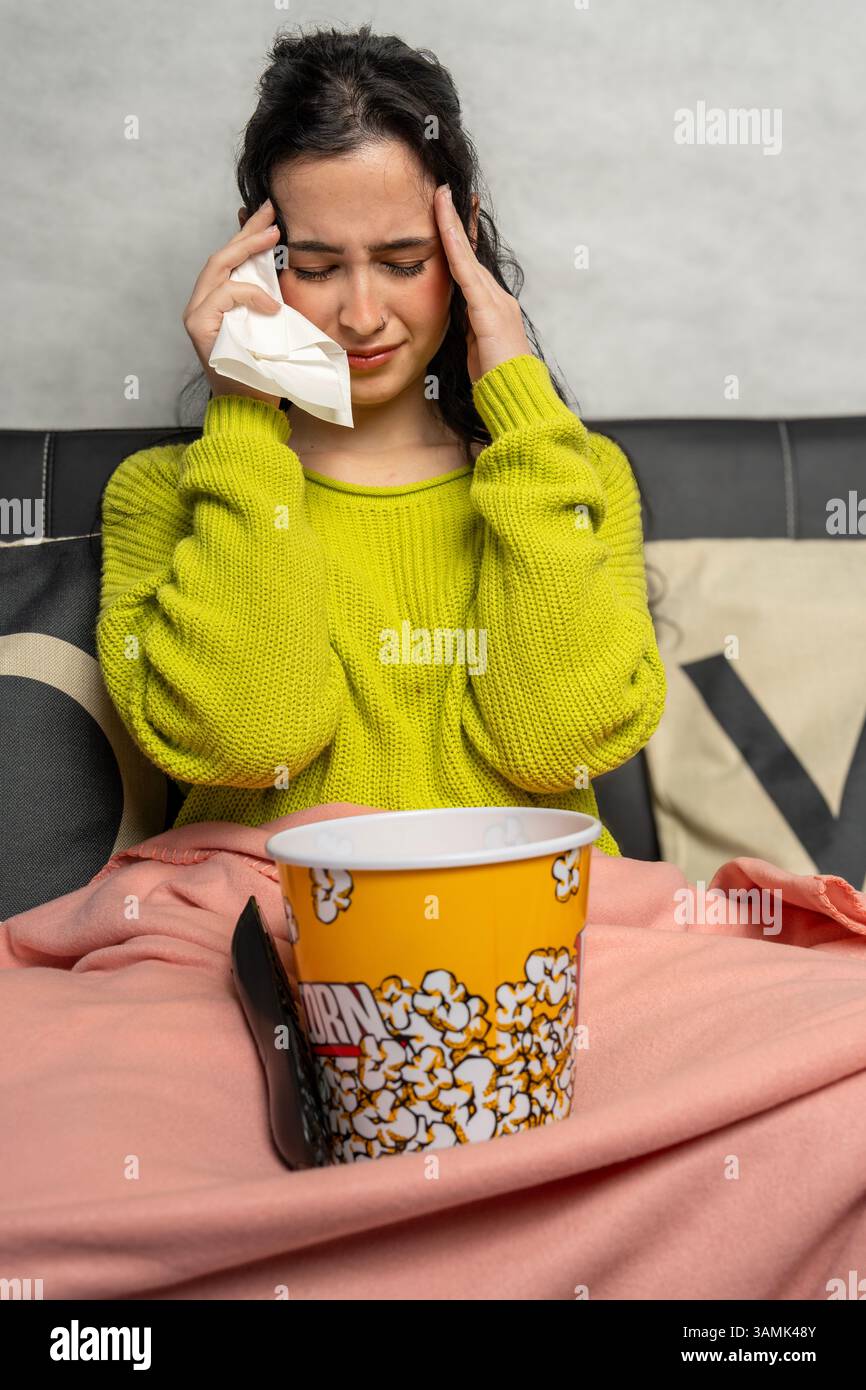 Traurige Frau, die sich einen Film ansieht und Tränen mit Taschentuch abwischt, während sie mit Popcorn-Eimer auf dem Sofa sitzt Stockfoto
