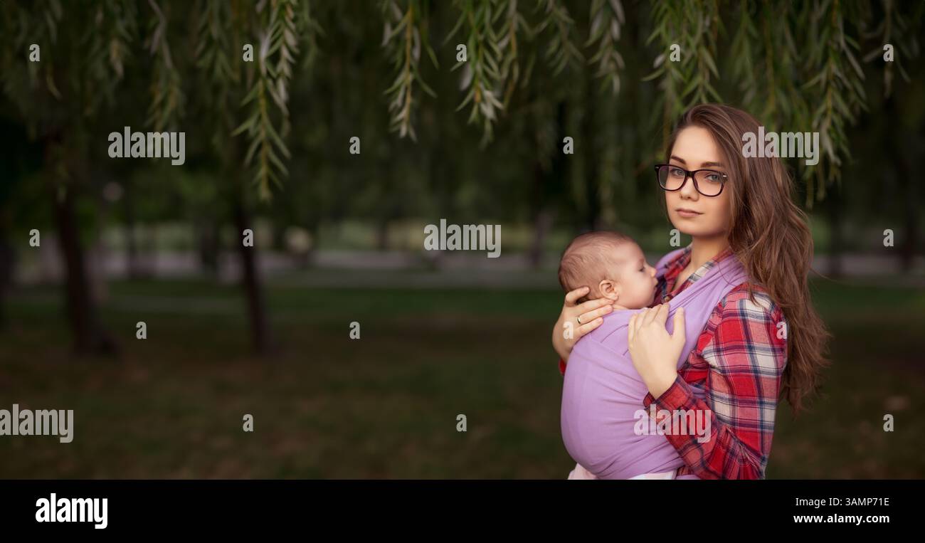 Junge Mutter mit Baby in Schlinge unter Weidenbaum im Park. Friedliche Bindung und Mutterschaft Stockfoto