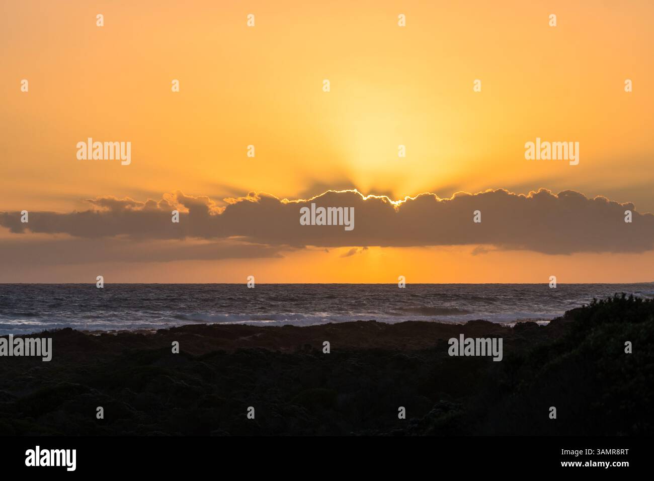Sonnenuntergang, Sonne und Strahlen hinter einer einzigen langen linearen Wolke in einem orangefarbenen Himmel, der den Horizont und das Meer oder den Ozean zeigt, im Vordergrund die dunkle Küstenlinie Stockfoto