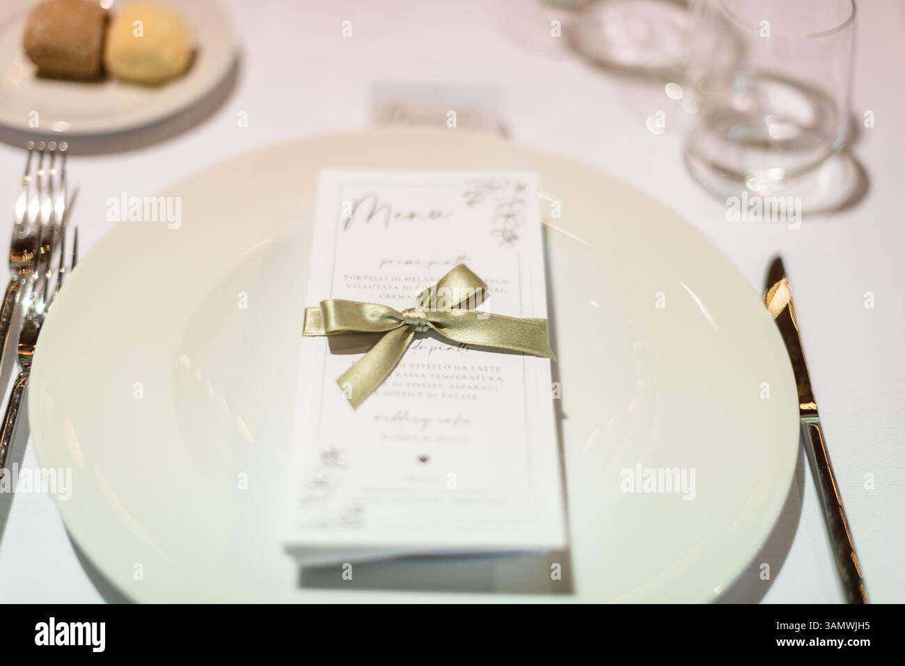 Ein wunderschön gestalteter Tisch verfügt über einen weißen Teller mit einem ordentlich gebundenen Menü und einem kleinen Dessert an der Seite, der für ein formelles Abendessen eingerichtet ist. Stockfoto