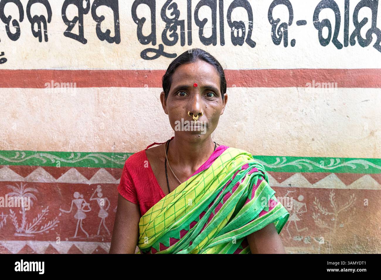 Porträt einer Frau mit Nasenringen und Schmuck, die vor einer bunten Schlammwand in Orissa, Indien, sitzt, indische Buchstaben an einer Wand Stockfoto