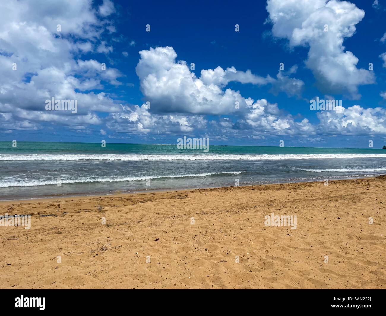 Ein perfekter Tag am Strand mit goldenem Sand, rollenden Wellen und einem hellblauen Himmel voller flauschiger Wolken. Schwimmer und Surfer genießen die Erfrischung - Smartphone-aufgenommenes Stockfoto