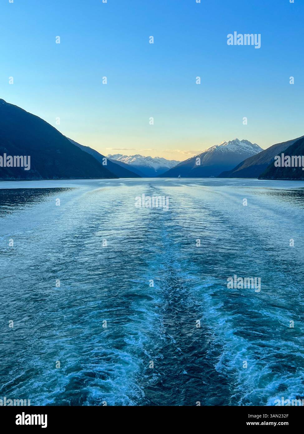 Ein atemberaubender Blick auf die Fjorde Alaskas bei Sonnenuntergang, der hinter einem Kreuzfahrtschiff eine ruhige Wache hinterlässt. Die schneebedeckten Berge umrahmen die Ruhe - Smartphone-aufgenommenes Stockfoto