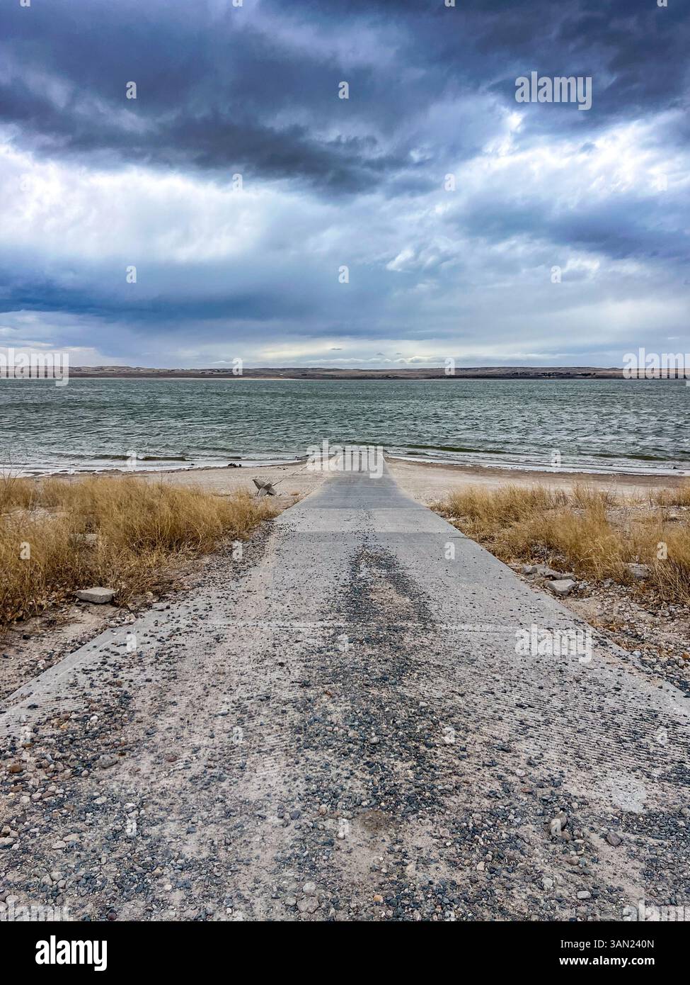 Eine zerklüftete Bootsrampe erstreckt sich unter schweren grauen Wolken in das abgehackte Seenwasser und fängt die Ruhe vor einem Sturm auf der offenen Prärie ein. - Smartphone-aufgenommenes Stockfoto