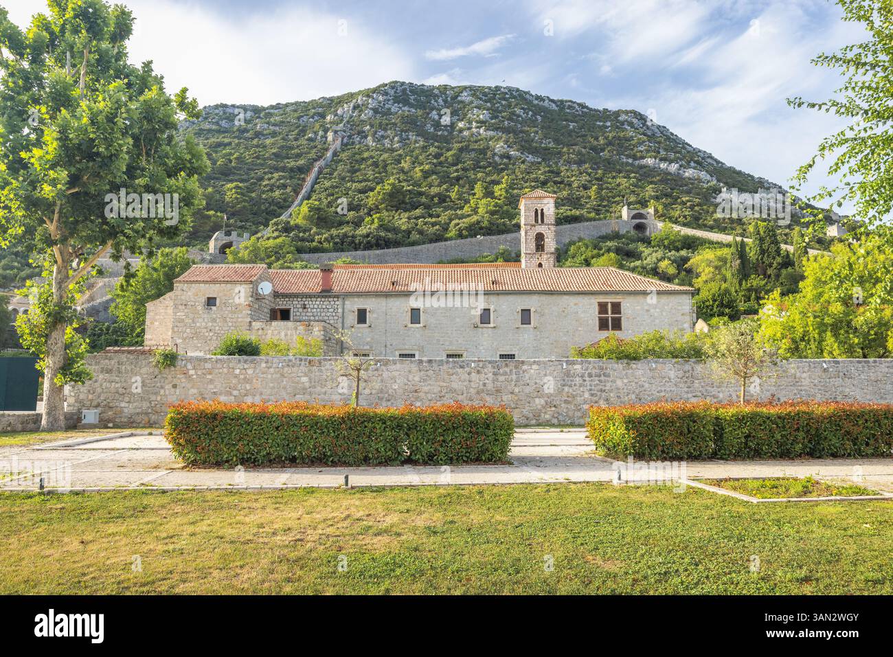 Ston, historische Stadt auf der Halbinsel Peljesac in Kroatien, Europa. Steingebäude mit einem Turm in der Nähe eines Hügels, der mit Bäumen bedeckt ist, und einem Weg, der zum t führt Stockfoto