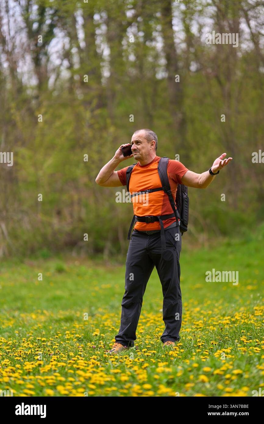 Wanderer in orangefarbenem Hemd, der am Telefon mit frustriertem Ausdruck spricht, während er auf einer blühenden Frühlingswiese steht Stockfoto