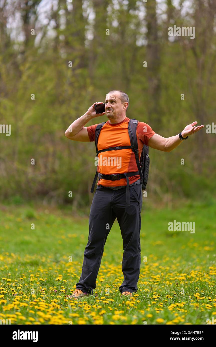Wanderer in orangefarbenem Hemd, der am Telefon mit frustriertem Ausdruck spricht, während er auf einer blühenden Frühlingswiese steht Stockfoto