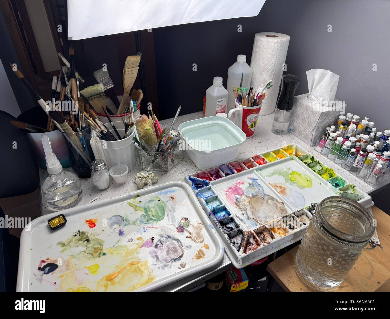 Beistelltisch neben dem Künstlerpult mit Aquarellzubehör. Palette, Farben, Pinsel, Mischschale und Wasserbehälter. Softbox-Studioleuchte. USA. - Smartphone-aufgenommenes Stockfoto