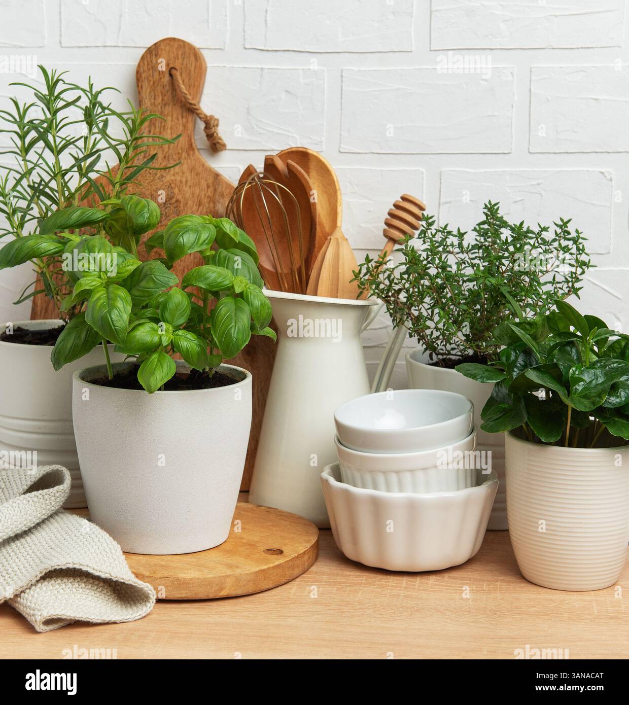 Aromatische Kräuter gedeihen in Töpfen neben Küchenutensilien und weißem Geschirr und verkörpern eine gemütliche Küchenatmosphäre vor einer weißen Ziegelwand Stockfoto