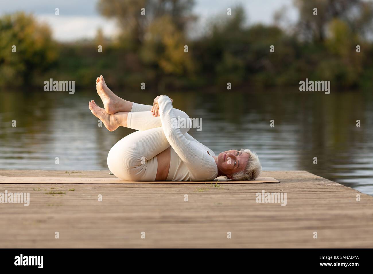 Eine Erwachsene Frau macht Yoga auf einem hölzernen Pier am Fluss, Pavanamuktasana posiert mit zwei Beinen, Windentspannung Stockfoto