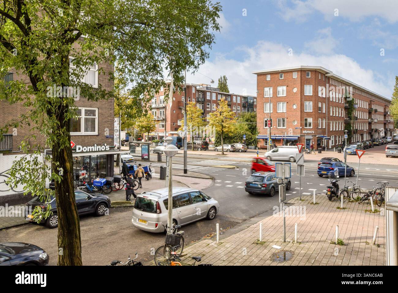 Eine lebhafte urbane Szene, die eine geschäftige Kreuzung mit Geschäften, Fahrzeugen und Fußgängern unter klarem blauen Himmel darstellt. Stockfoto