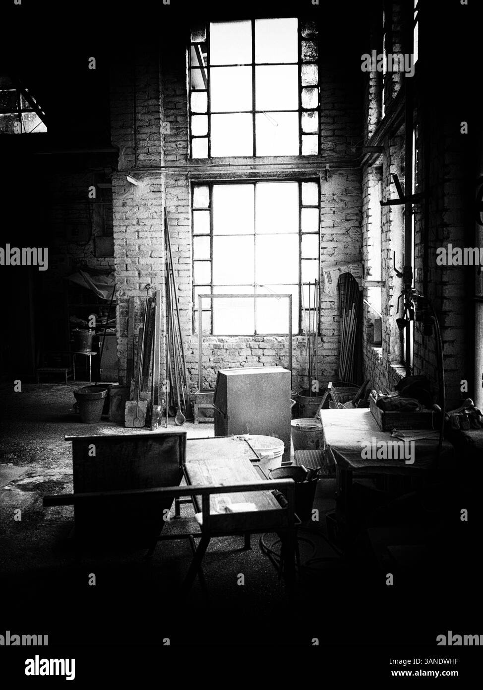 Ein Schwarzweißfoto eines alten Glasfabrikinnenraums mit großen Industriefenstern und verstreuten Werkzeugen. Stockfoto