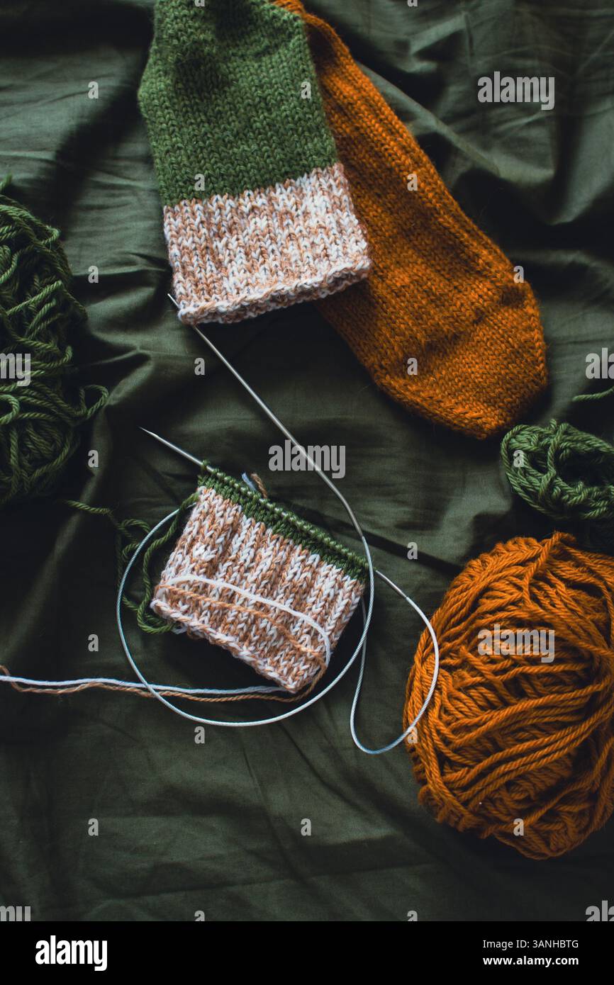 Dunkelgrüner und brauner Garnball mit gestrickten Socken und Stricknadeln. Wollfäden mit tiefgrünem Hintergrund. Gewebte Häkelnaht. Strickhobbyarbeit Stockfoto
