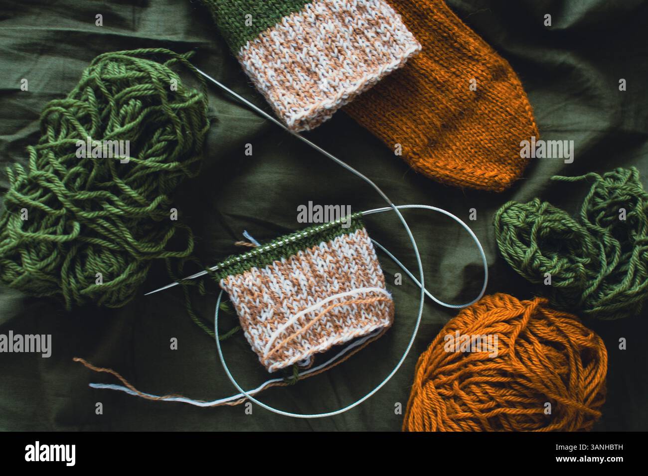 Dunkelgrüner und brauner Garnball mit gestrickten Socken und Stricknadeln. Wollfäden mit tiefgrünem Hintergrund. Gewebte Häkelnaht. Strickhobbyarbeit Stockfoto