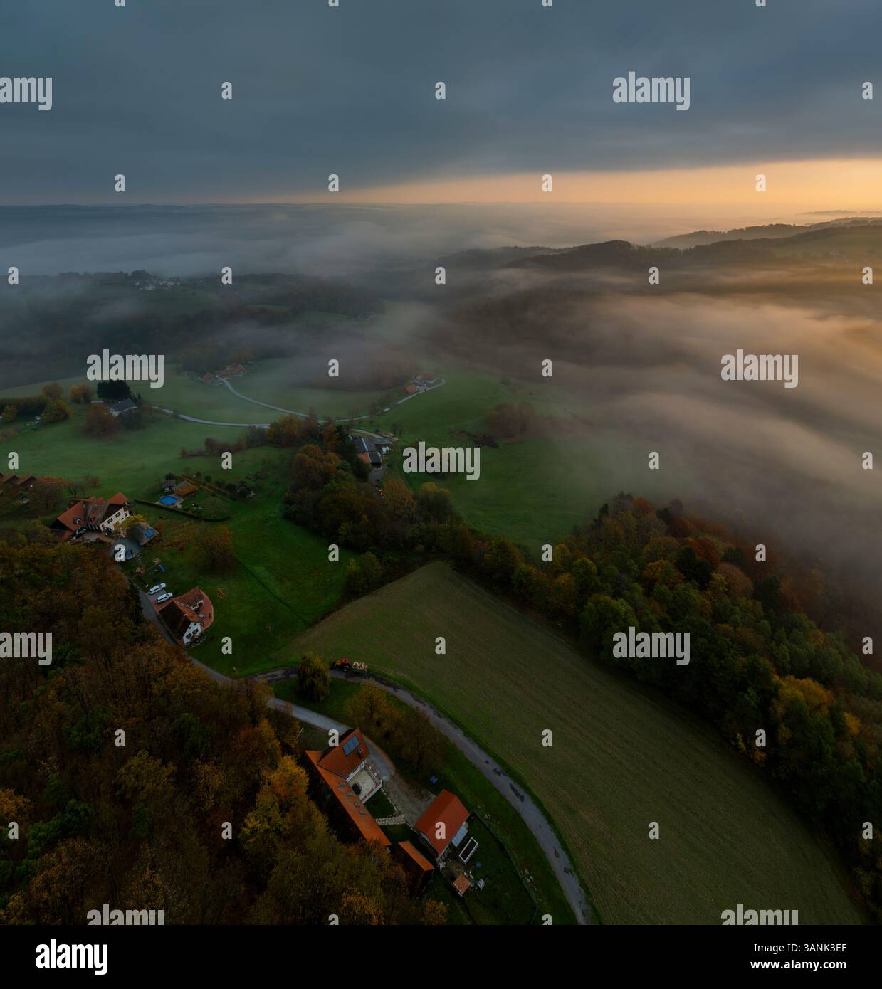 Aus der Vogelperspektive auf nebeligen Sonnenaufgang über dichtem Wald und grünem Tal mit Häusern und Feldern, Bad Loipersdorf, Österreich. Stockfoto