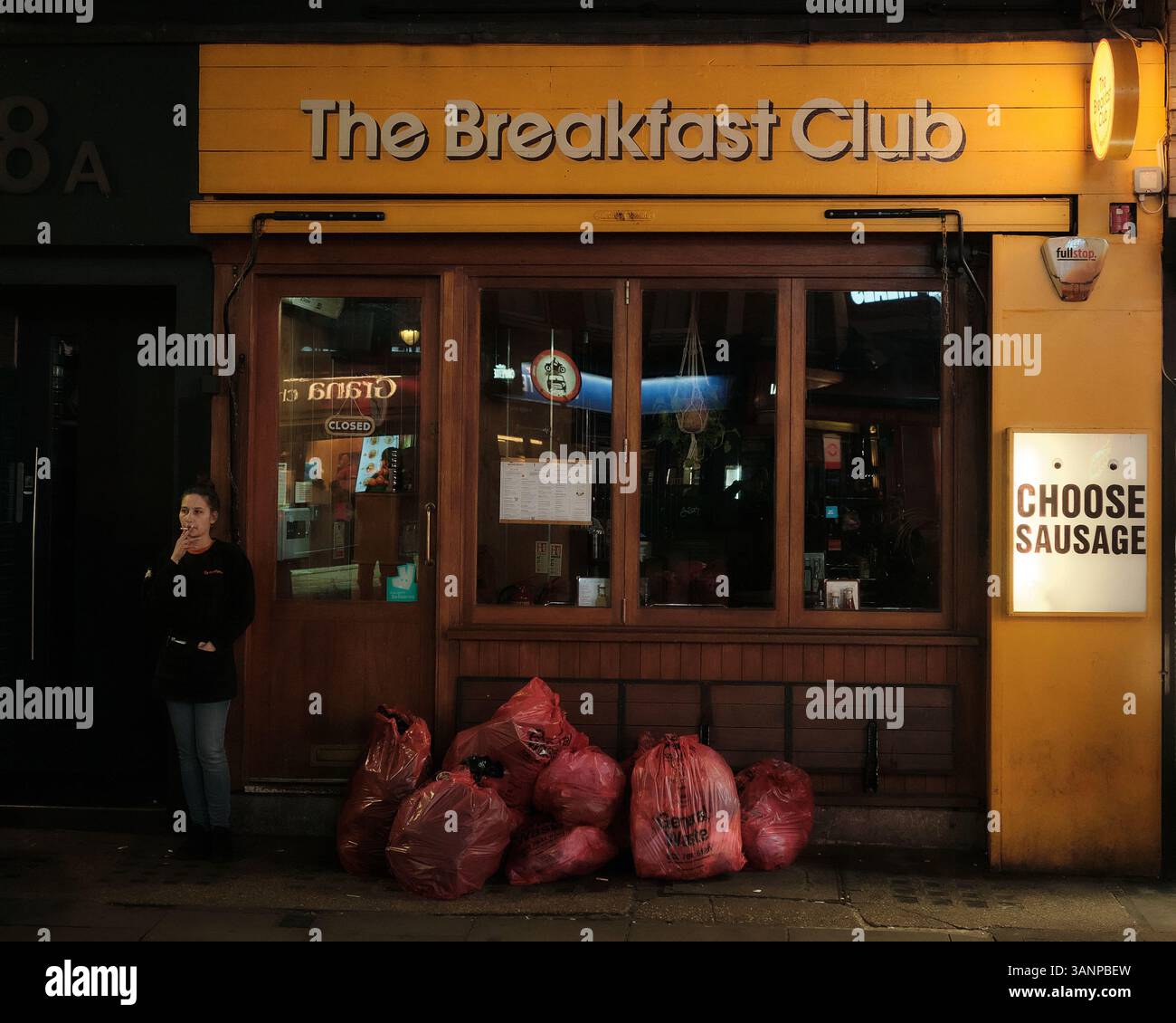 Eine Frau raucht nachts in Soho, London, vor einem geschlossenen Café, neben pinkfarbenen Mülltüten. Stockfoto