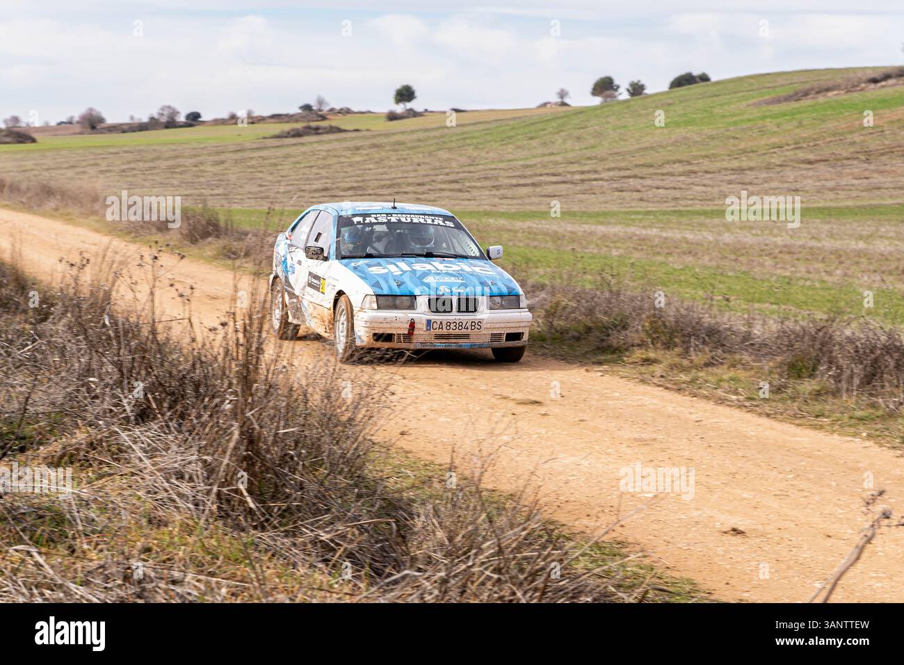 Madrid, Spanien; 15.02.2025: Ein blau-weißer BMW 323i E36 fährt auf einer Kies-Rallye-Strecke und schleudert Staub, während er durch die Landschaft fährt Stockfoto