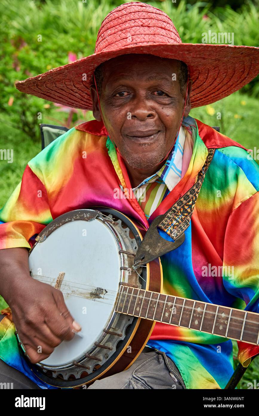Nahaufnahme-Porträt eines älteren karibischen Volksmusikers, der einen roten Strohhut und ein farbenfrohes Hemd trägt und im Freien Banjo spielt. Stockfoto