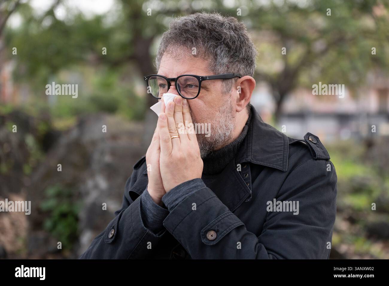 Mann mittleren Alters niesen mit einem Gewebe in einer natürlichen Umgebung, zeigt Symptome einer saisonalen Allergie oder Erkältung. Begriff der Atemgesundheit. Stockfoto