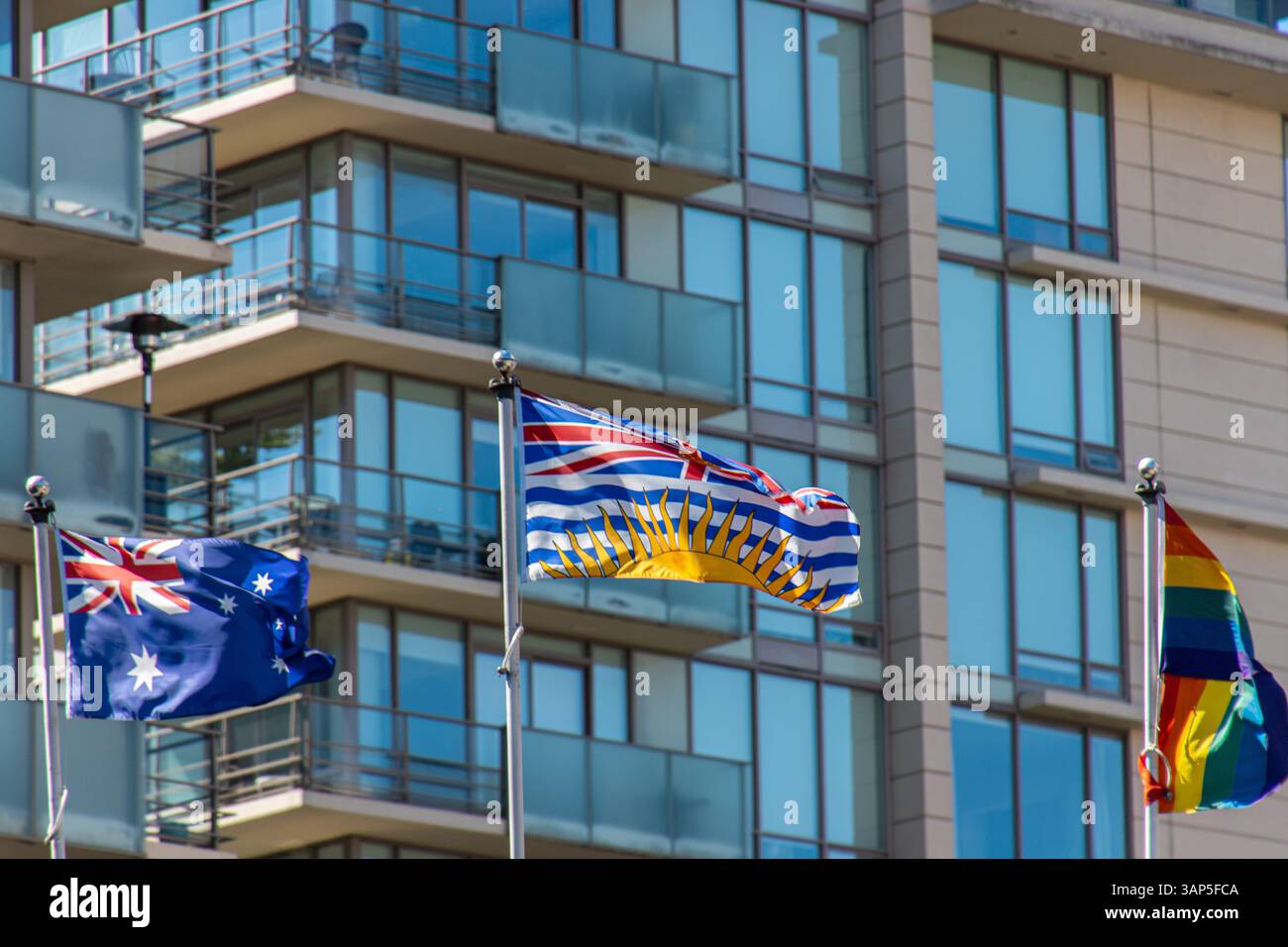 Die Flagge von British Columbia winkt deutlich mit australischen und Pride-Flaggen im Hintergrund vor einem modernen Glaswohnhaus. Stockfoto