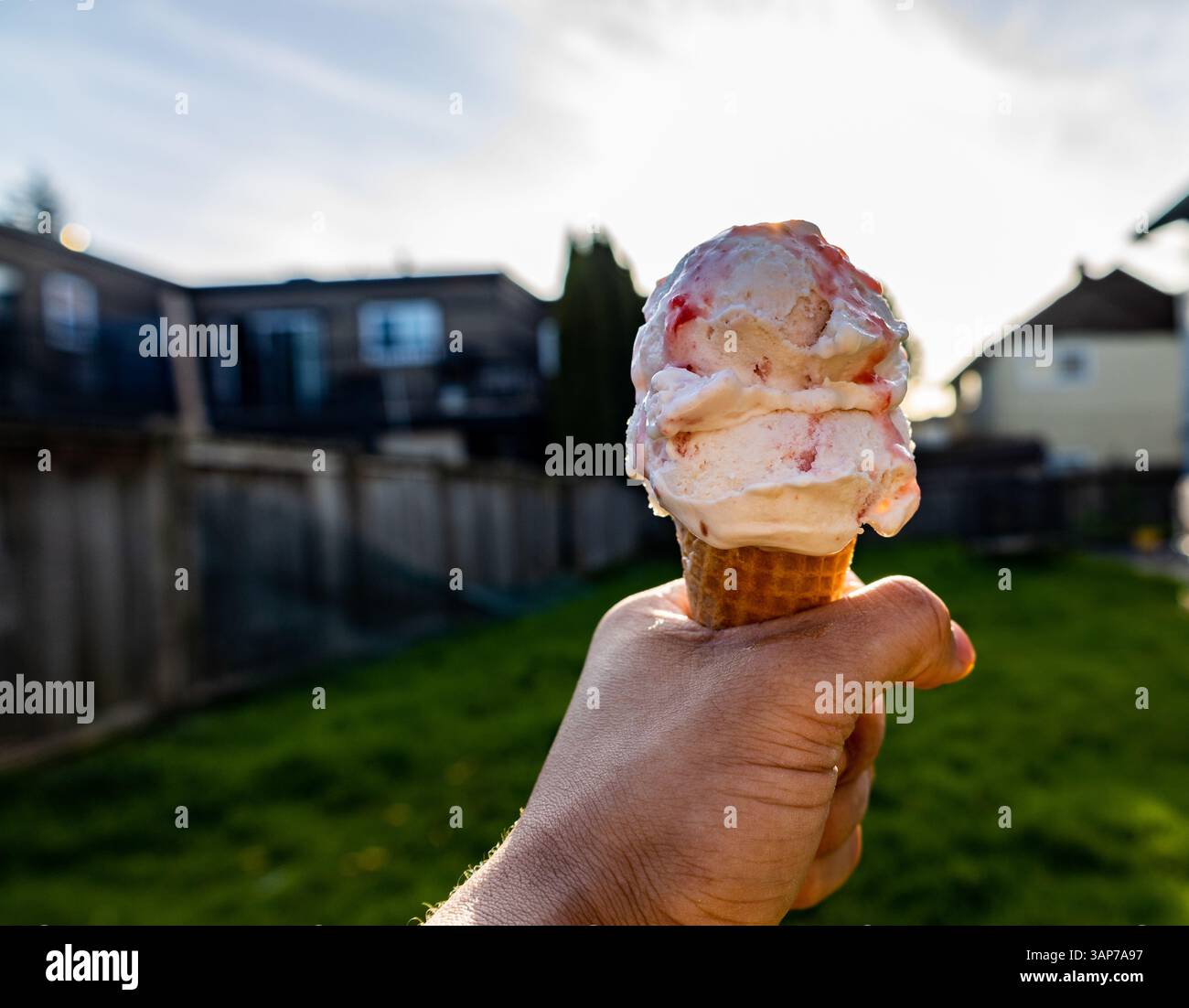 Die Hand hält Erdbeereiskegel im Hinterhof mit der Sonne hinter dem Kegel Stockfoto