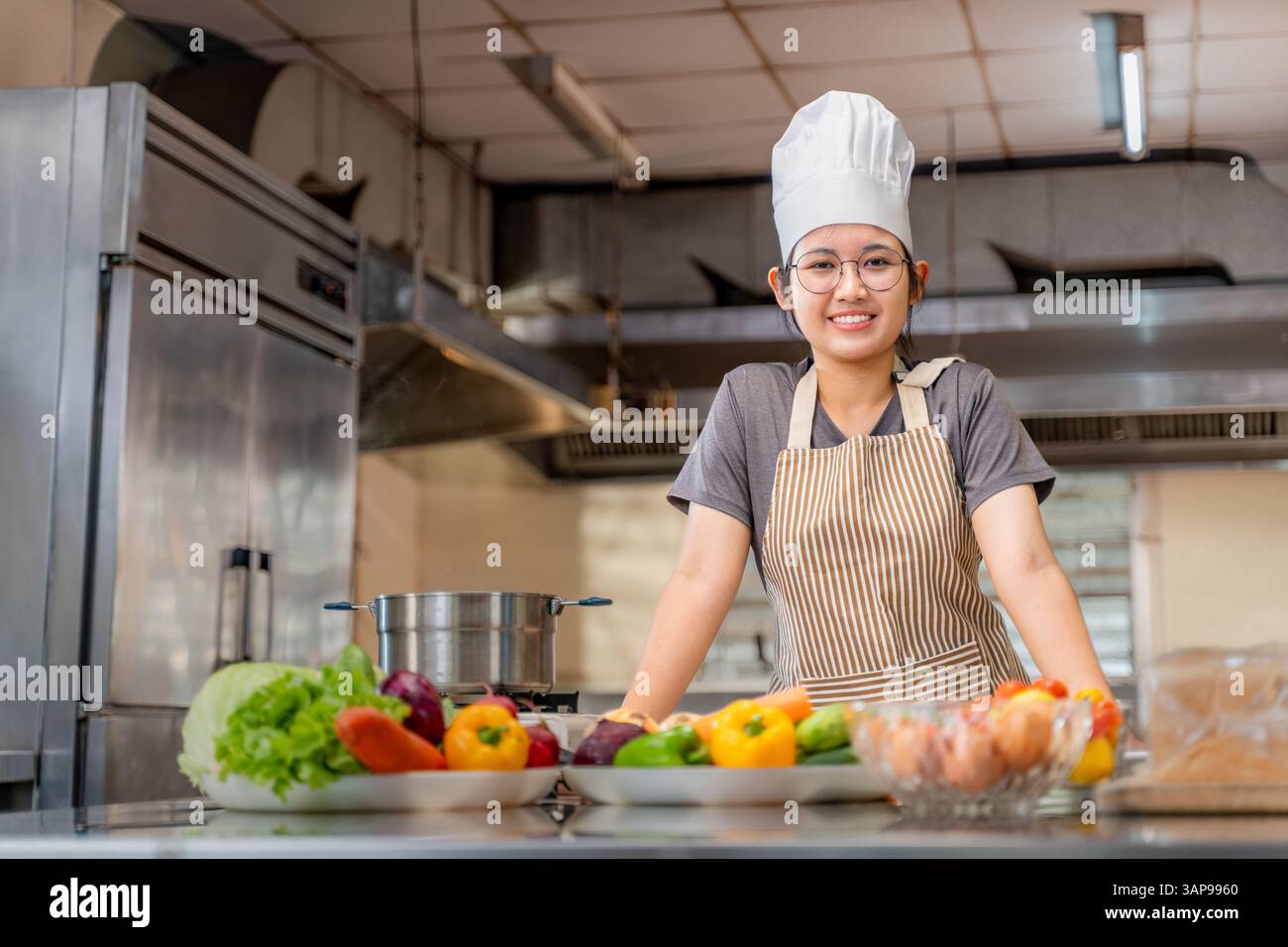Porträt einer jungen Küchenchefin in einer professionellen Küche, die hinter einer Theke steht, gefüllt mit frischem Gemüse und Kochzutaten. Tragen eines s Stockfoto