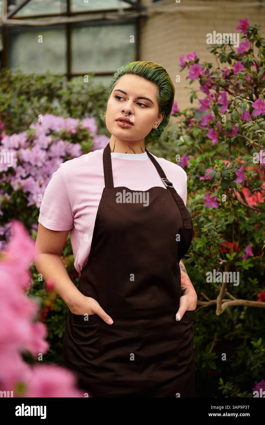 In einem Gewächshaus konzentriert sich eine selbstbewusste junge Frau mit grünen Haaren auf ihre Arbeit unter Blumen. Stockfoto