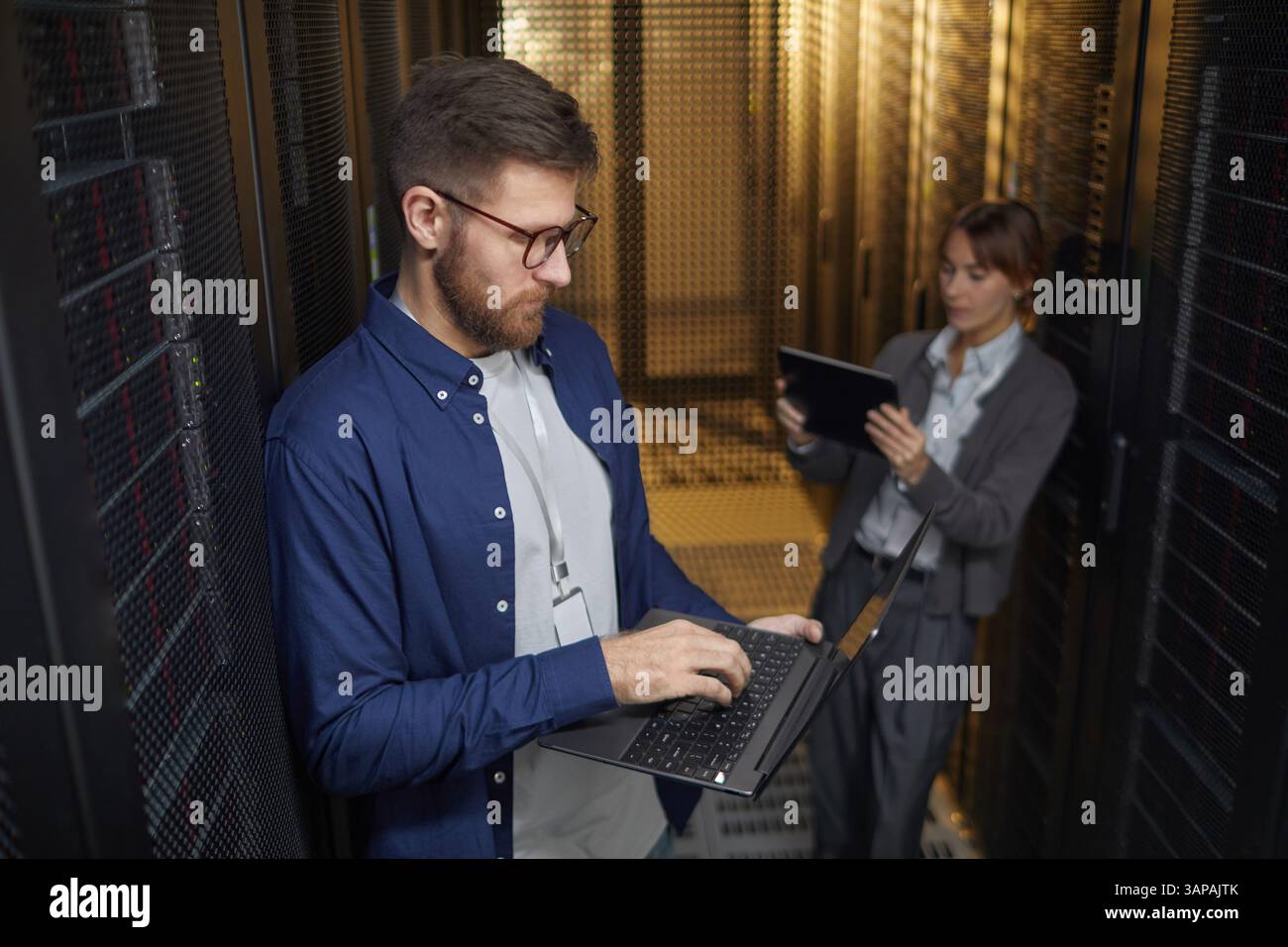 Zwei IT-Experten, die Servergeräte in modernen Rechenzentren verwalten. Eine Person verwendet ein Notebook, während eine andere ein Tablet verwendet, um sicherzustellen, dass alle Systeme betriebsbereit sind Stockfoto