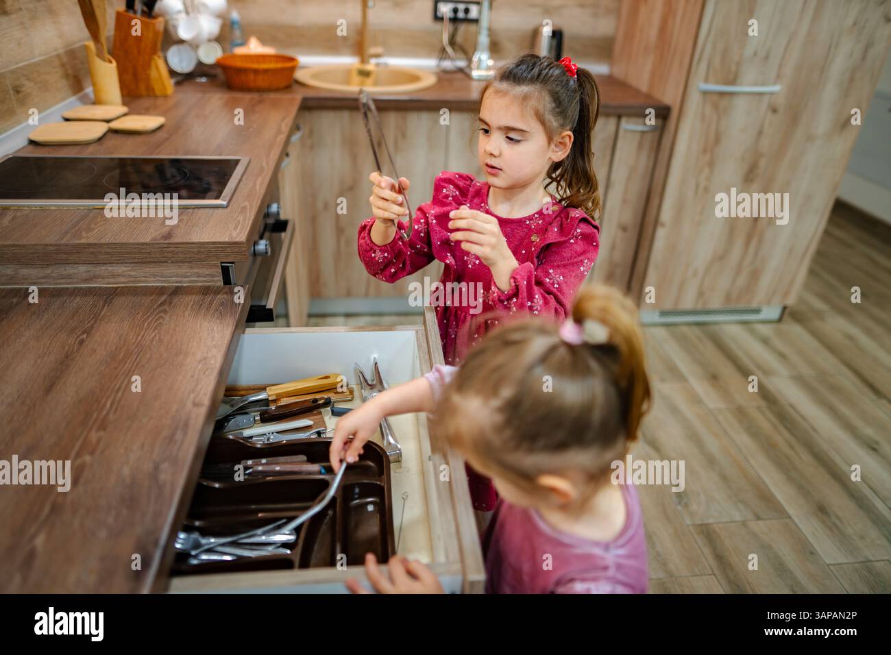 In einer wunderschön gestalteten Küche untersuchen zwei Mädchen eine Schublade, die mit Utensilien überflutet ist. Ihre Neugier weckt Lachen und Aufregung, während sie sich entschlüsseln Stockfoto