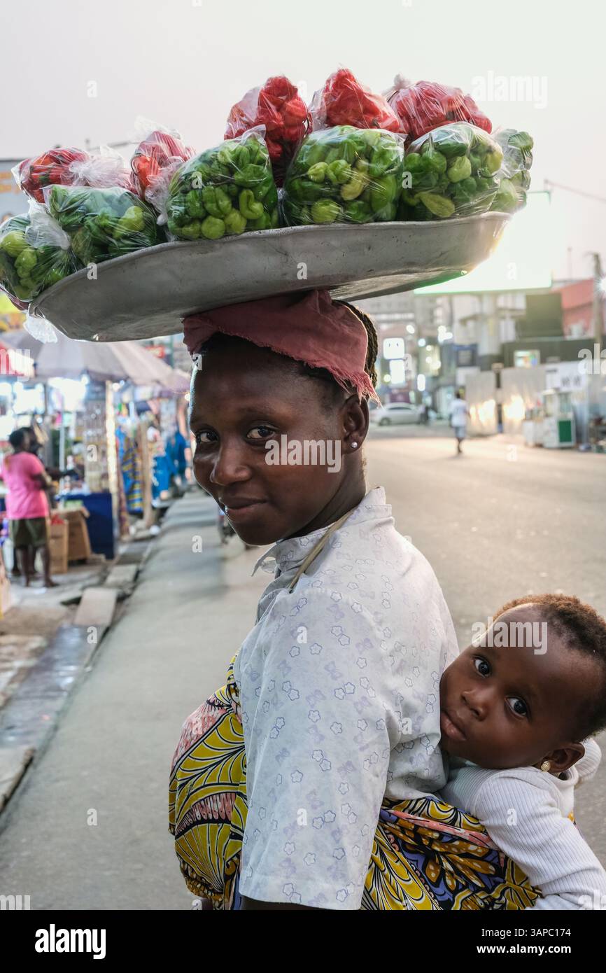 Accra, Ghana. Oxford Street Scene. Frau trägt Baby auf dem Rücken, Tablett mit Paprika in Taschen auf dem Kopf. Stockfoto