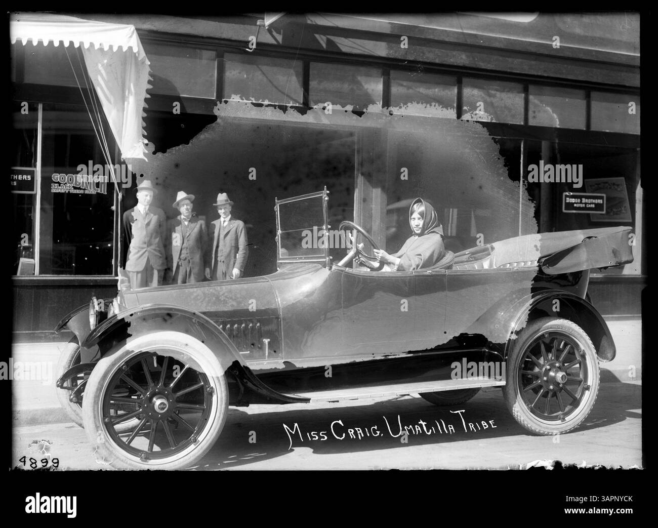 Foto von Miss Craig, einer Amerikanerin aus dem Umatilla-Stamm, die hinter dem Steuer eines Autos sitzt und drei Männer in Anzügen in der Nähe stehen. Das Gebäude wirbt für Dodge Brothers Motor Cars. Stockfoto