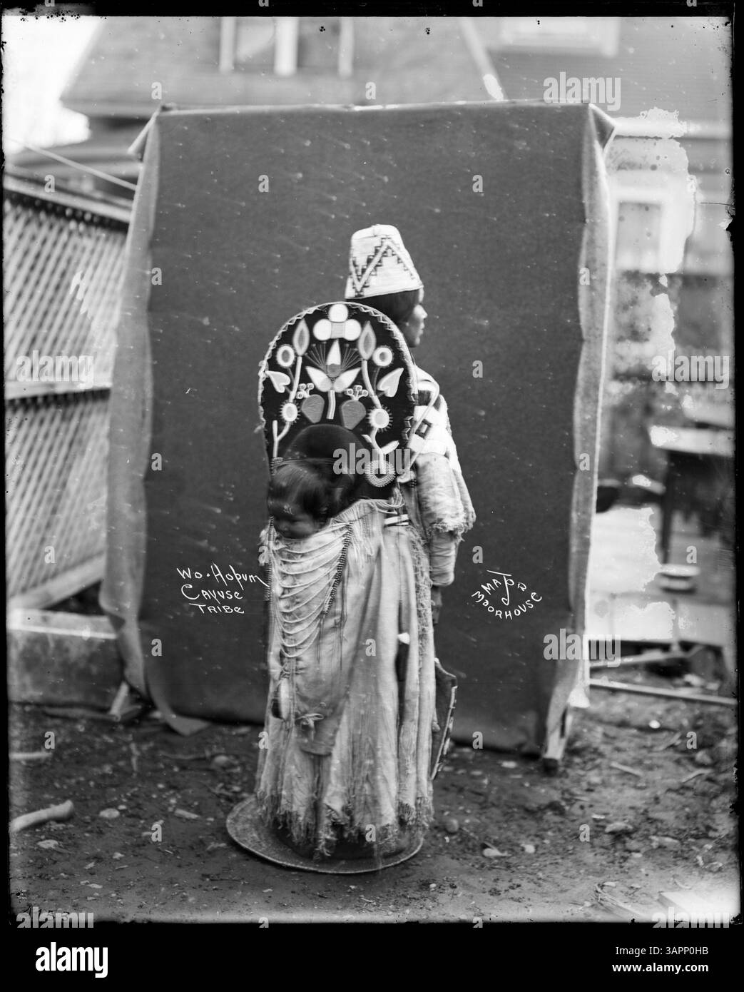 Dieses Foto zeigt Wo-ho-Pum, einen Cayuse-Indianer, gekleidet in Insignien, der ein Kind in einer Wiege hält. Das Bild ist Teil der Lee Moorhouse Kollektion und zeigt die indianische Kultur und traditionelle Kleidung. Stockfoto