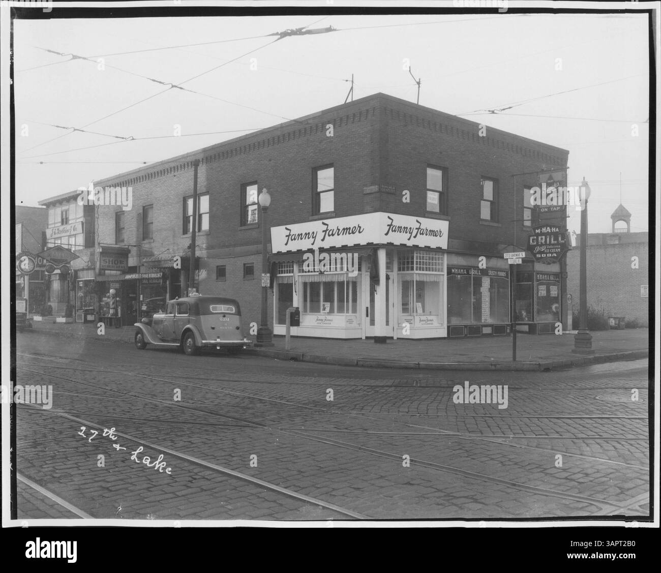 Dieses historische Bild zeigt verschiedene Unternehmen entlang der Lake Street mit einem gelben Taxi, das vor einem Grill geparkt ist. Eine Waage ist außerhalb des Gebäudes zu sehen, und Bahngleise sind in der Nähe zu sehen. Stockfoto