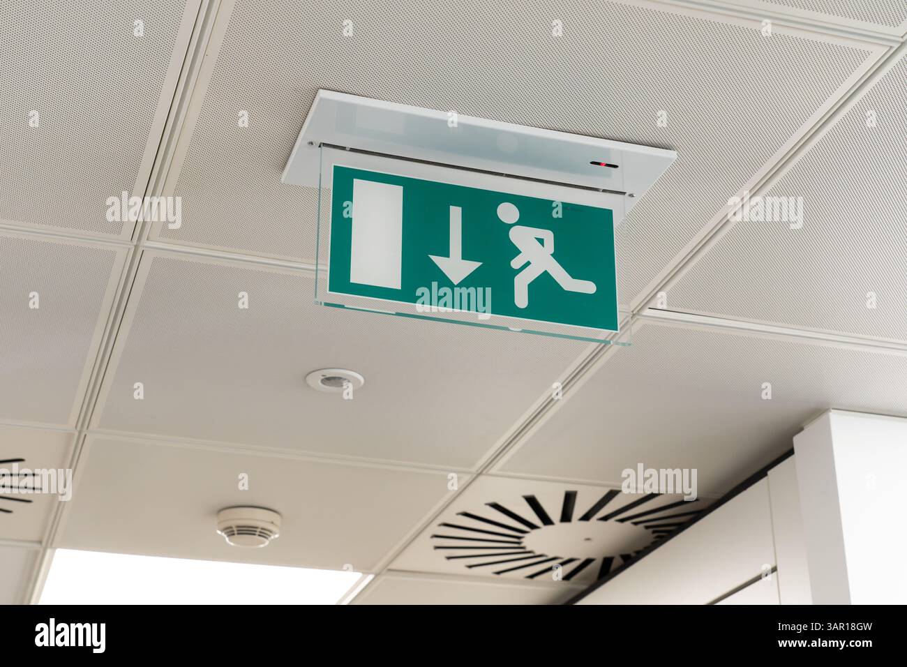 An der Decke montiertes LED-Notausgangsschild mit Pfeil, der in Richtung der Brandschutztür in einem britischen Büro zeigt, mit Rauchmelder dahinter. Konzept: Brandschutz Stockfoto