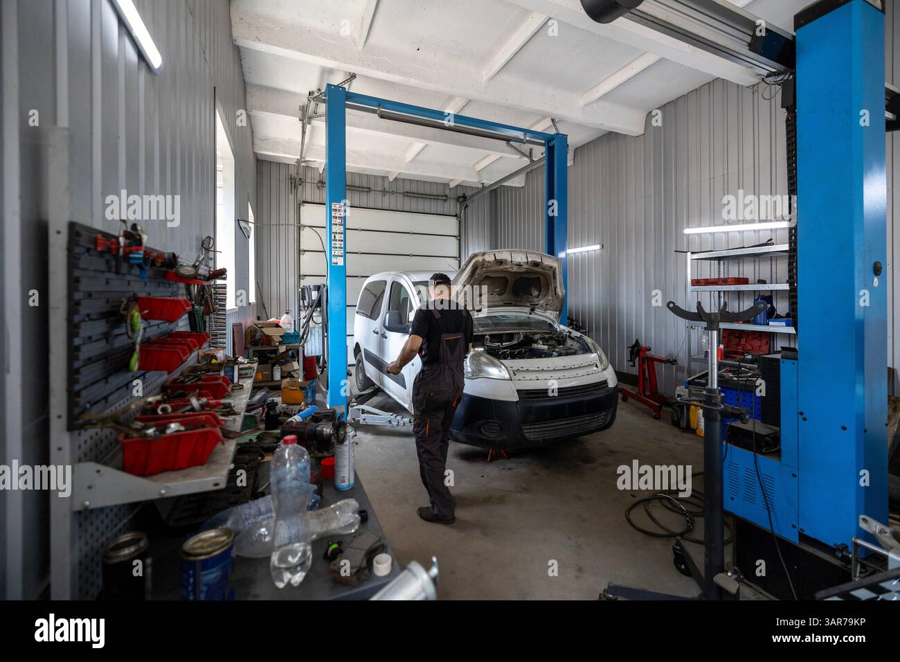 Ein Mechaniker arbeitet an einem weißen Transporter mit offener Motorhaube in einer geschäftigen Autowerkstatt, die mit einem Hebebühne und verschiedenen Werkzeugen ausgestattet ist Stockfoto
