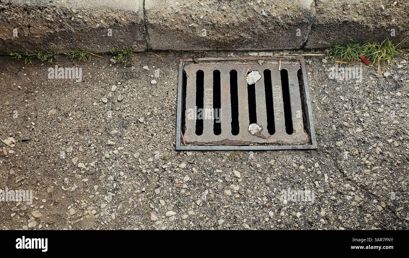 Nahaufnahme eines rostigen Metallgitters, der einen Sturmabfluss auf einer Schotterstraße bedeckt und Anzeichen von Verschleiß zeigt Stockfoto