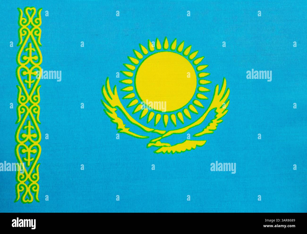 Die Flagge hat einen hellblauen Hintergrund, eine goldene Sonne und einen Adler im Flug. Auf der linken Seite befinden sich komplizierte gelbe Muster, die die Nation repräsentieren Stockfoto