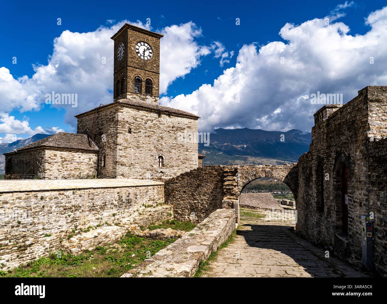 Ein historischer Uhrenturm aus Stein steht vor dem Hintergrund dramatischer Wolken und Berge. Die Szene zeigt alte Steinmauern und einen Pfad Stockfoto