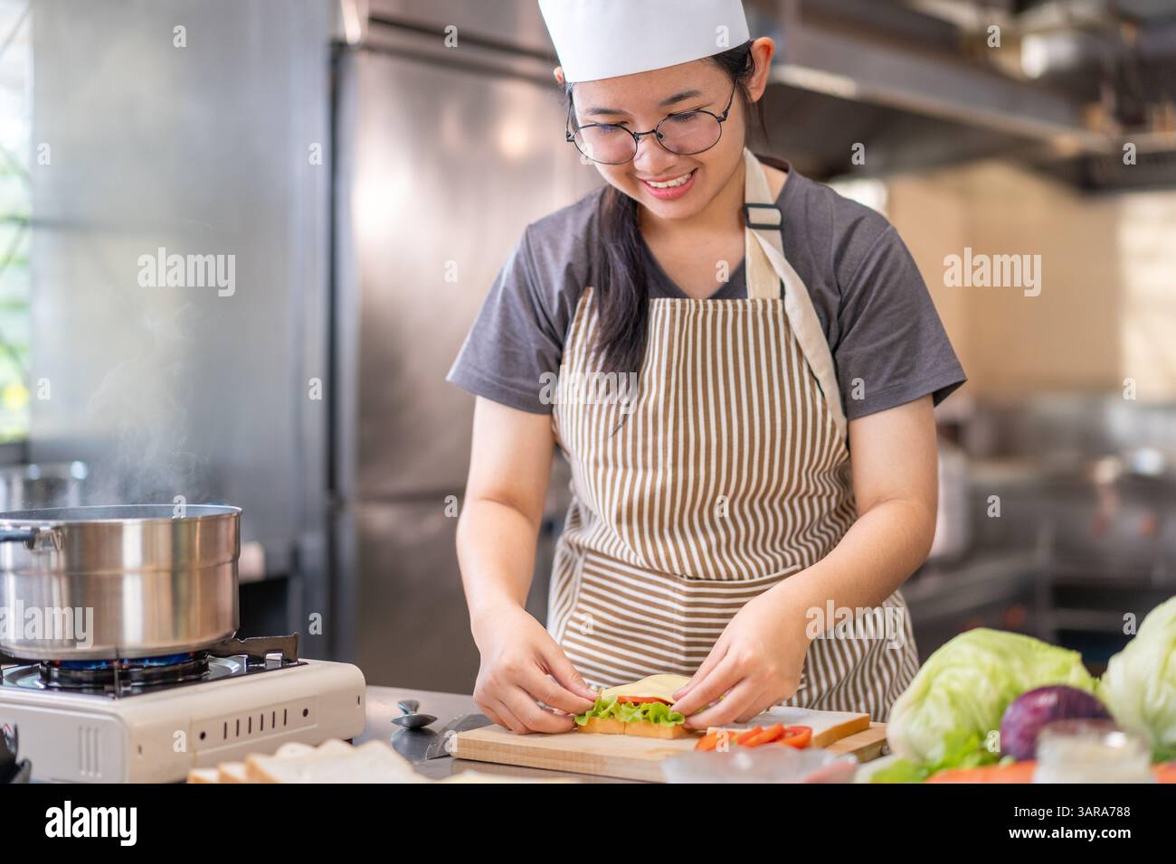 Eine fröhliche junge Küchenchefin, die eine gestreifte Schürze und einen weißen Kochhut trägt, sieht man, wie sie ein frisches Sandwich in einer kommerziellen Küche zubereitet. Dieses Bild vermittelt Stockfoto