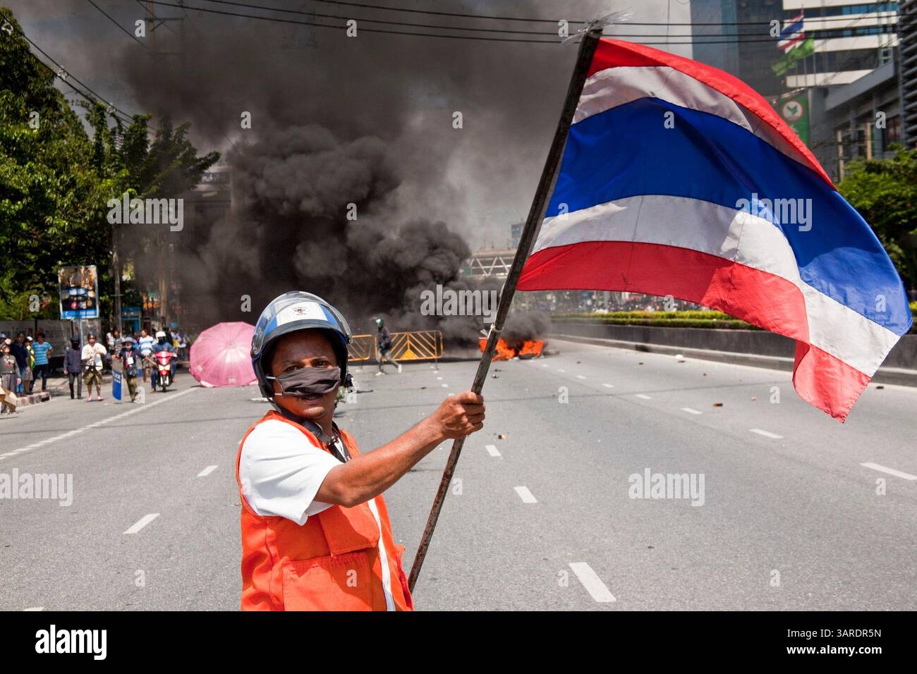 14. Mai 2010 - Bangkok, Thailand - ein regierungsfeindlicher Demonstrant schwingt die thailändische Flagge vor einer brennenden Barrikade auf der Rama IV Rd Die thailändischen Sicherheitskräfte haben Live-Patronen abgefeuert, nachdem sie ein schwer verteidigtes Demonstrantenlager in Bangkok wegen Zusammenstößen, bei denen zwei Menschen getötet wurden, versiegeln wollten. Botschaften wurden geschlossen, als Demonstranten einen Polizeibus in Brand setzten und Feuerwerke auf Truppen schossen, die auch mit Tränengas und Gummigeschossen reagierten. Die Demonstranten wollen, dass der Premierminister zurücktritt und Wahlen ausruft. Viele der sogenannten Red-Shirt-Demonstranten unterstützen den ehemaligen Premierminister Thaksin Shinawatra, der sich mit dem „Roten-Shirt-Protest“ beschäftigt Stockfoto