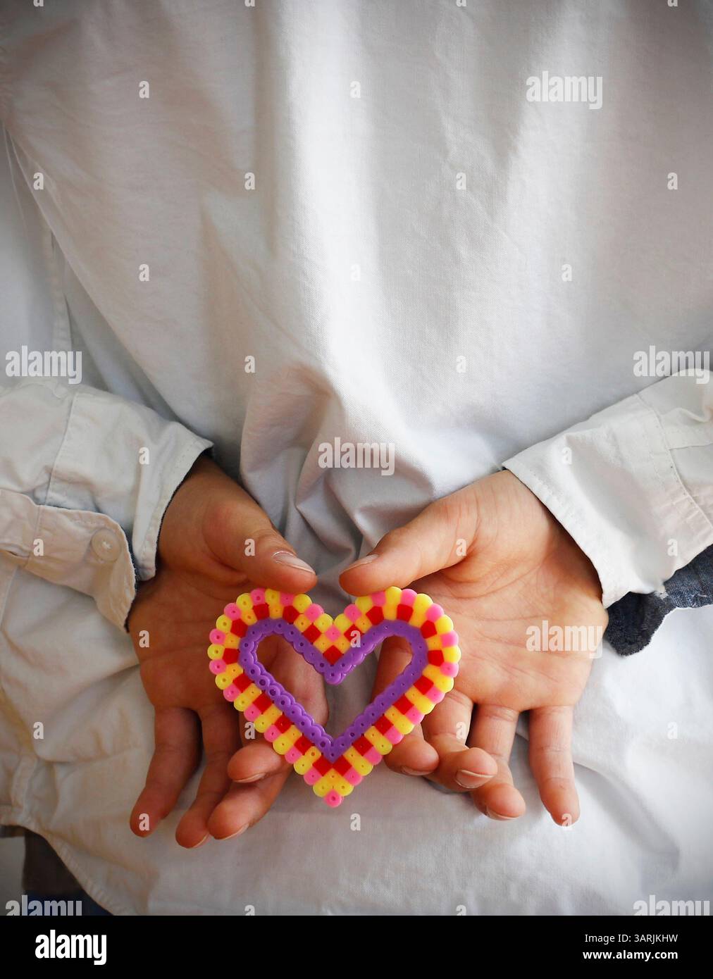 Die Hände des kaukasischen Jungen halten ein buntes Plastikherz aus Bügelperlen hinter seinem Rücken. Muttertag- und Vatertag-Konzept mit Kopierraum. Stockfoto