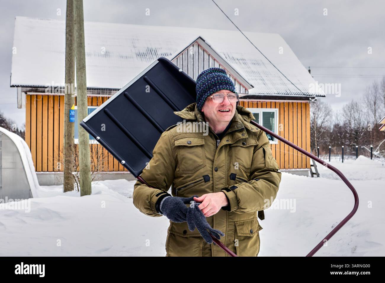 Bei routinemäßigen Arbeiten im Freien mit Schneeräumung sieht ein älterer, erwachsener Mann in seinen 50ern ernsthaft mit Schaufel neben einem ländlichen Haus aus, umgeben von tiefem Schnee. Stockfoto