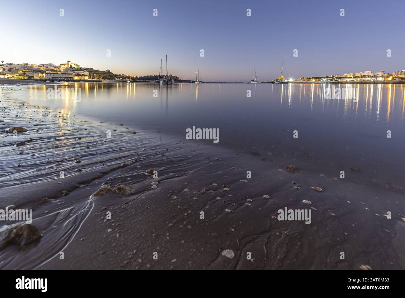 Nachtaufnahme von einem Sandstrand und Hafen bei Ebbe. Panoramabild eines beleuchteten historischen Stadtzentrums bei Sonnenaufgang in Ferragudo, Portimao, Algarve, Stockfoto